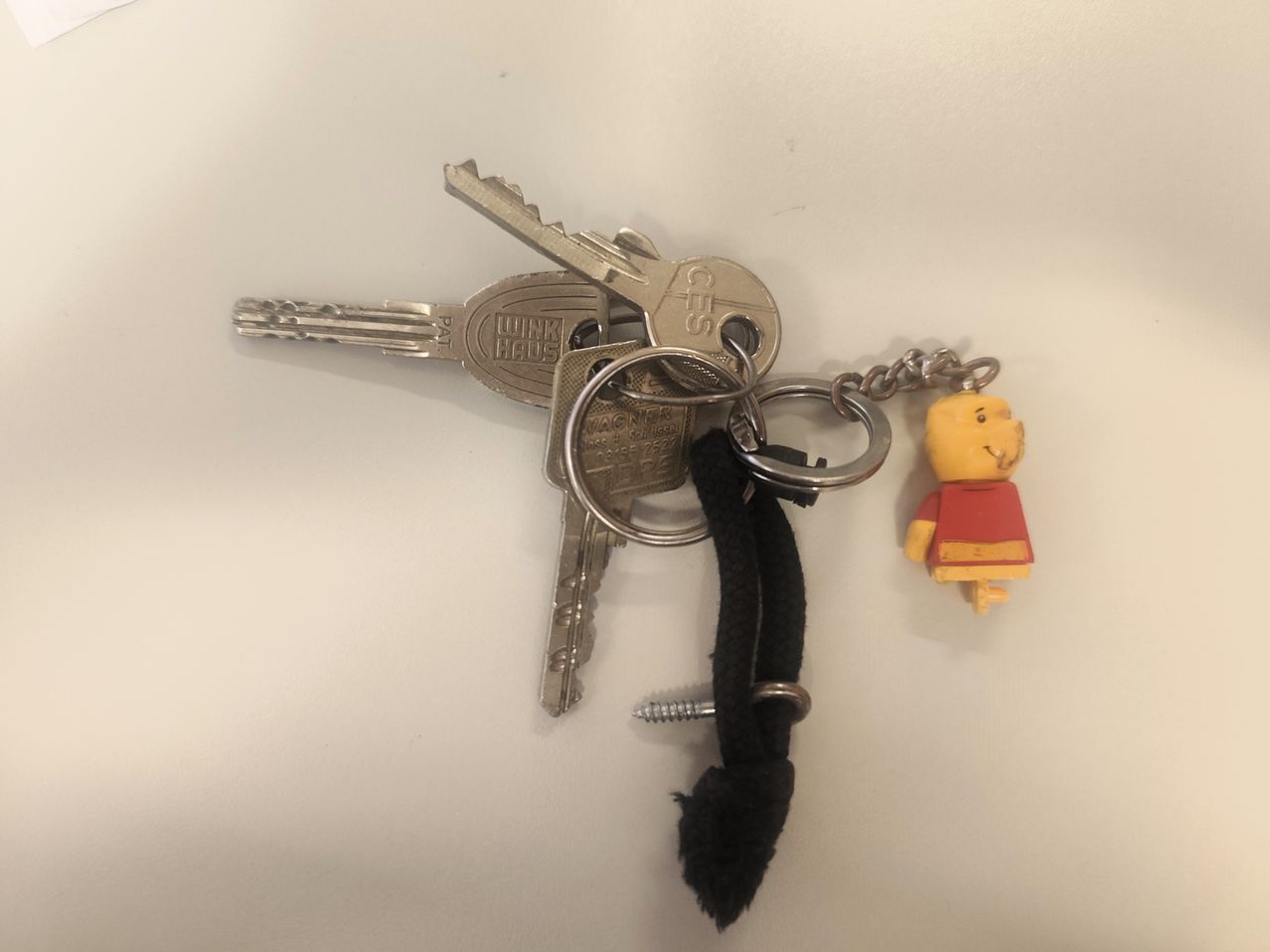 Zwei Schlüssel, eine Schraube und ein Winnie-the-Pooh-Schlüsselanhänger auf einer weißen Oberfläche. Die Schlüssel haben das Wort WINKHAUS darauf.