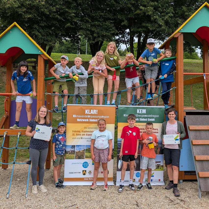 Eine Gruppe von Kindern und Erwachsenen posiert für ein Foto in einem Park. Sie stehen um einen Spielplatz herum mit Schildern, die 'KLARI' und 'KEM' lesen. Die Kinder halten Urkunden. Bäume und Gras sind im Hintergrund.