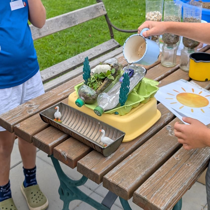Zwei Kinder spielen mit einem wissenschaftlichen Experiment auf einem Holztisch. Die Einrichtung besteht aus Plastikflaschen und einer gelben Ablage mit Pflanzen und einer kleinen Entenfigur.