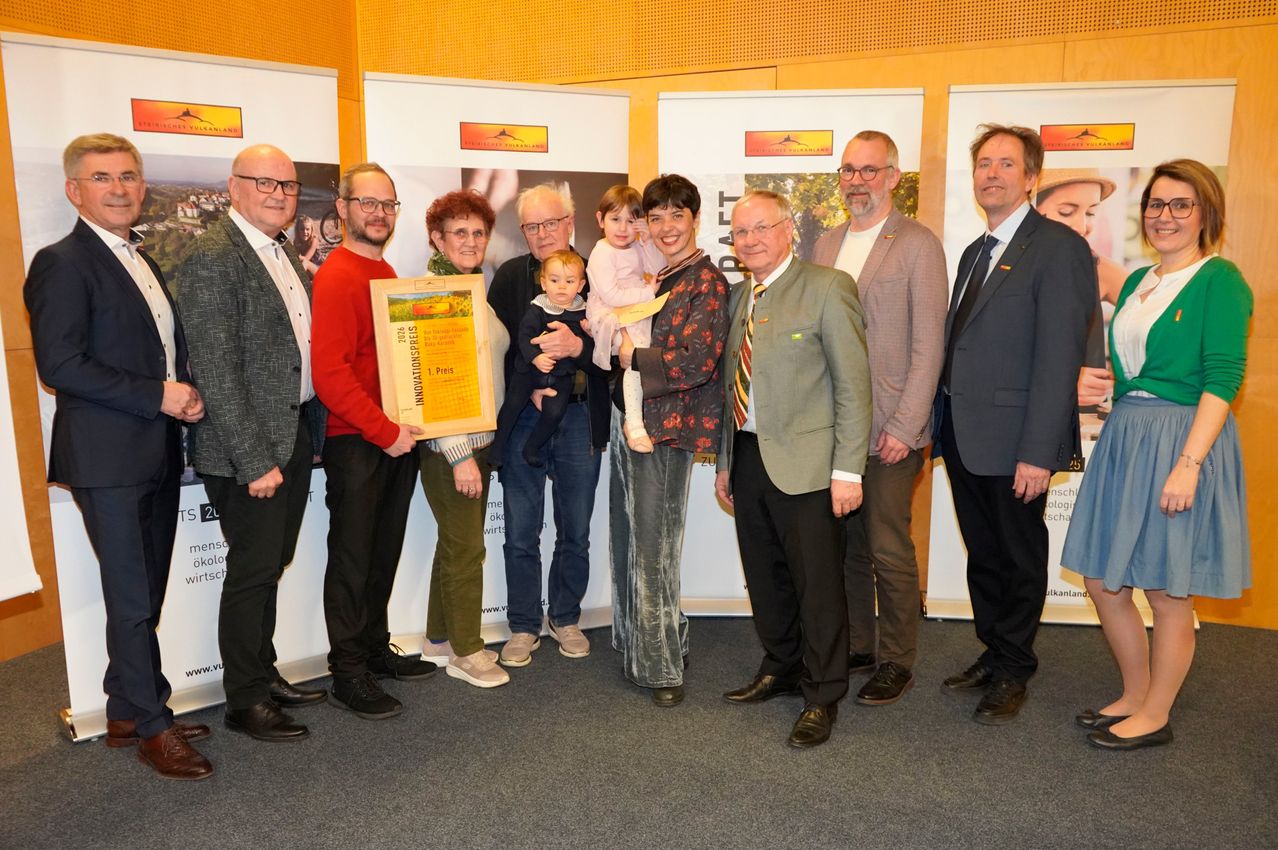 Eine Gruppe von Menschen steht vor Bannern, hält einen gerahmten Preis und lächelt. Einige halten Kinder, während andere zusehen. Die Szene erscheint formell und feiertlich.