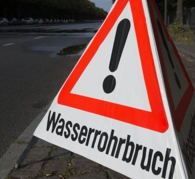 Ein dreieckiges Warnschild am Straßenrand weist mit einem roten Dreieck und einem schwarzen Ausrufezeichen auf einen Wasserrohrbruch hin.