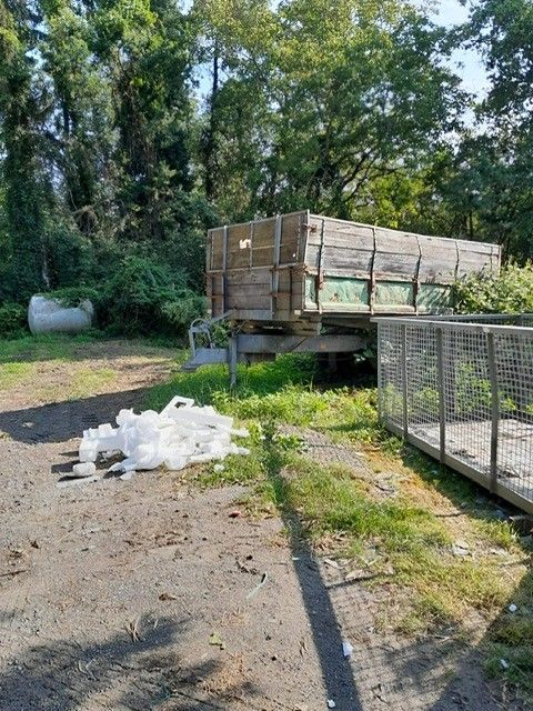 Ein alter Holzanhänger steht auf einem Feldweg mit einem Haufen Plastikabfällen in der Nähe. Ein Metallkäfig befindet sich rechts, und ein großer Tank steht hinter dem Anhänger.