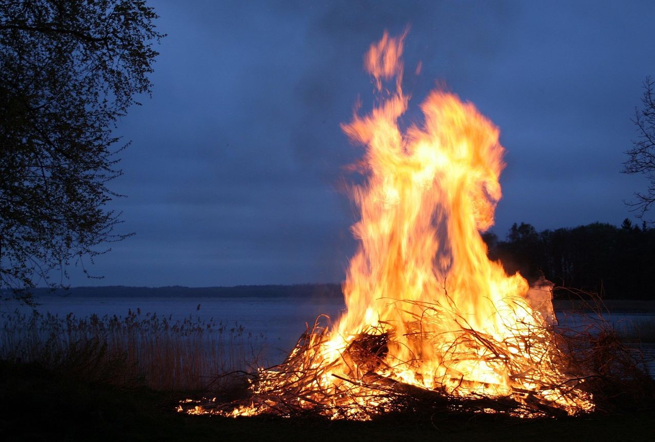 Ein Lagerfeuer brennt hell in der Nähe eines Sees bei Nacht, mit hochreichenden Flammen und Bäumen und einer ruhigen Wasserfläche im Hintergrund.