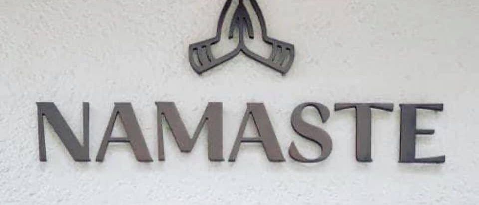 Ein metallisches Schild mit dem Wort Namaste in fetten, großgeschriebenen Buchstaben über einem stilisierten Handsymbol. Das Schild ist an einer weißen Wand befestigt.