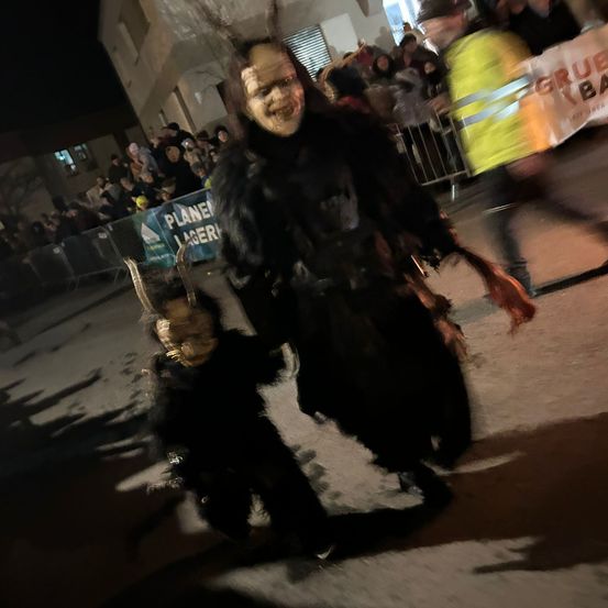 Ein Mann und ein Kind, beide in schwarzen Kostümen gekleidet, gehen nachts durch eine Menschenmenge. Der Mann trägt eine Maske und hält eine Waffe. Dahinter sind ein Sicherheitsbeamter und ein Banner zu sehen.