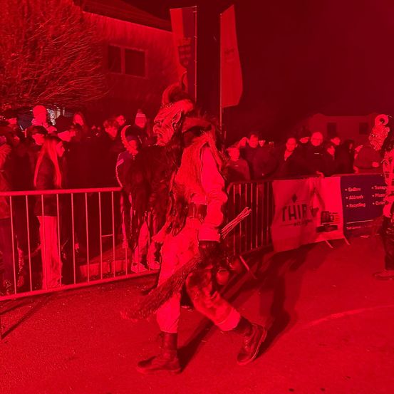 Eine Person in einem Kostüm geht bei roter Beleuchtung bei Nacht an einer Menge vorbei. Dahinter befindet sich ein Zaun und ein Banner.