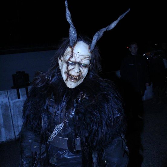 Eine Person in einer gehörnten Maske und Fellkostüm steht in einem dunklen Bereich, möglicherweise bei einer Halloween-Veranstaltung. Dahinter ist ein Mann in einem schwarzen Jackett und mit Brille teilweise zu sehen.