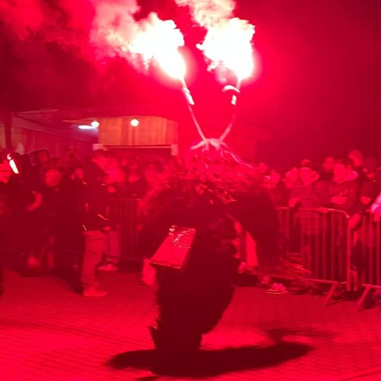 Ein Künstler steht inmitten einer rot beleuchteten Darbietung, mit zwei Fackeln, die Feuer aussenden, umgeben von einem Publikum auf einem gepflasterten Boden.