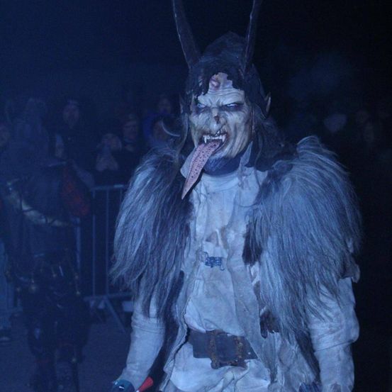 Eine Person in einem Wolfskostüm mit einer langen heraushängenden Zunge steht vor einer Menge in einem dunklen Bereich.