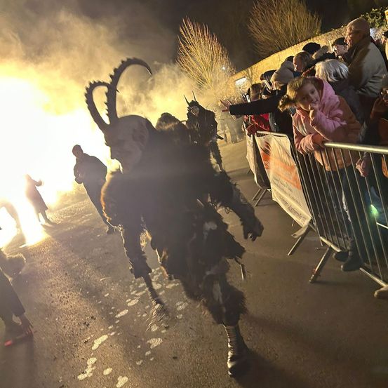 Eine Menschenmenge beobachtet einen Mann in einem Hörnerkostüm, der bei Nacht eine Straße entlang in Richtung eines Feuers geht.
