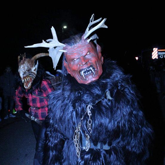 Zwei Personen in Kostümen, eine trägt eine rote Maske und Geweih, die andere eine gehörnte Maske, stehen bei Nacht auf einer Straße. Im Hintergrund ist ein Schild mit der Aufschrift 'FEUER' zu sehen.