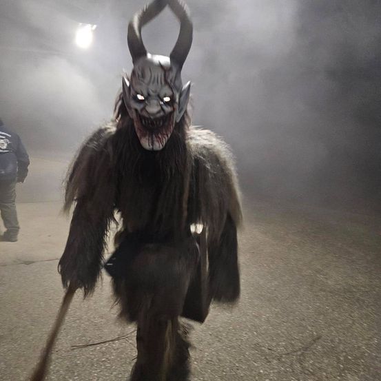 Eine Person in einem gruseligen Dämonenkostüm mit Hörnern und weißen Augen geht im Nebel. Eine Person in einer Jacke steht hinter ihr.