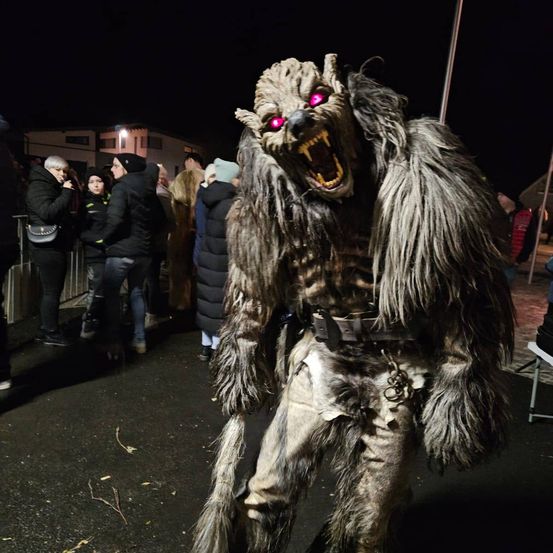Eine Person, die als Wolf verkleidet ist und leuchtend rote Augen hat, steht mitten in einer Menschenmenge bei Nacht. Die Menge scheint die Person zu beobachten, und einige halten eine Kamera.