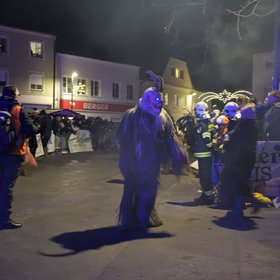Eine Menschenmenge beobachtet eine Person in einem Kostüm mit leuchtenden Augen auf einer Straße bei Nacht. Im Hintergrund sind Gebäude und eine Straßenlaterne zu sehen.