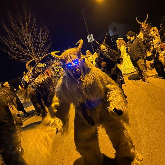 Eine nächtliche Szene mit Menschen in Kostümen, einer mit blauen Augen und Hörnern, die wahrscheinlich an einem Fest teilnehmen.