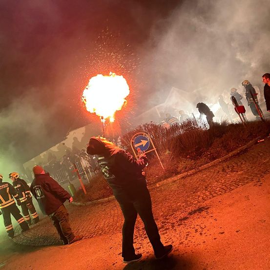 Ein großes Feuer brennt in der Nacht, mit einer Menge von Menschen hinter einer Barriere, einige in Feuerwehruniformen, und eine Person vorn, die auf eine Fackel bläst.