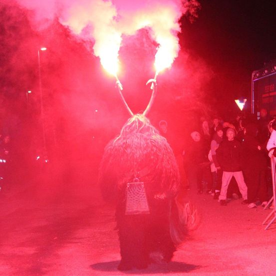 Eine Person in einem pelzigen Kostüm hält zwei flammende Fackeln vor einer Menschenmenge. Sie sind von rotem Rauch und Straßenlaternen umgeben.