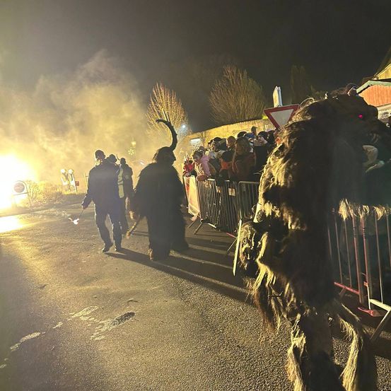 Eine Nachtveranstaltung mit Menschen in Kostümen, die auf einer Straße gehen, während andere hinter einer Barriere zusehen. In der Ferne brennt ein Feuer.