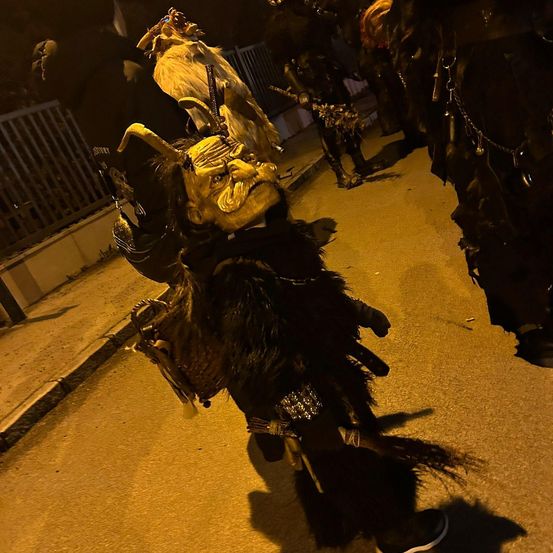 Eine Person in einem Kostüm mit einer großen Maske geht nachts eine Straße entlang, mit anderen Personen in Kostümen dahinter. Der Bereich ist schwach beleuchtet.