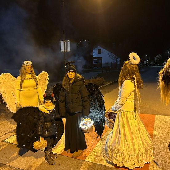 Vier Personen in Halloween-Kostümen stehen nachts auf einer Straße. Zwei Frauen in Engelskostümen, ein Kind in Teufelskostüm und eine Frau in Hexenkostüm.