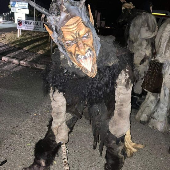 Eine Person in einem gehörnten Kostüm steht bei Nacht im Freien mit anderen, in der Nähe eines Schildes, das 'Heimlauf' steht.
