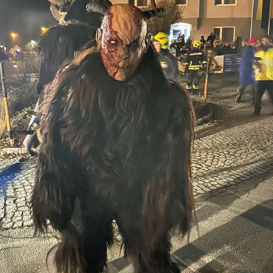 Eine Person in einem pelzigen Dämonenkostüm steht nachts auf einer Kopfsteinpflasterstraße, mit Feuerwehrleuten und Zuschauern im Hintergrund.
