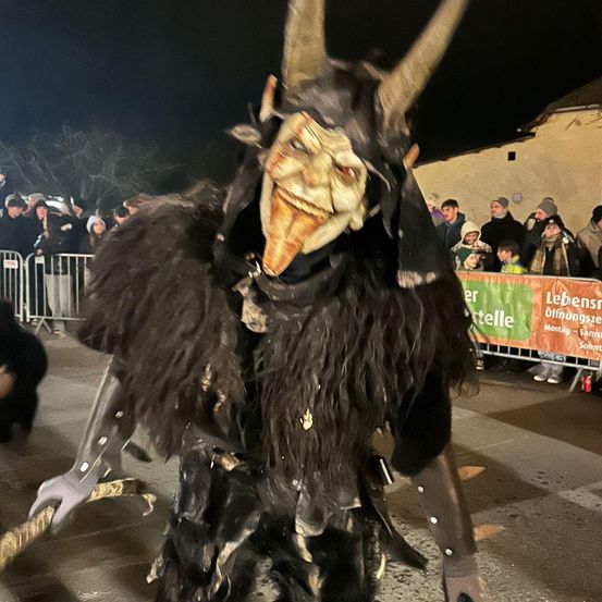 Eine Person als Teufel mit Hörnern verkleidet, der in einer Parade geht, mit einer Menschenmenge, die hinter einer Barriere zuschaut.