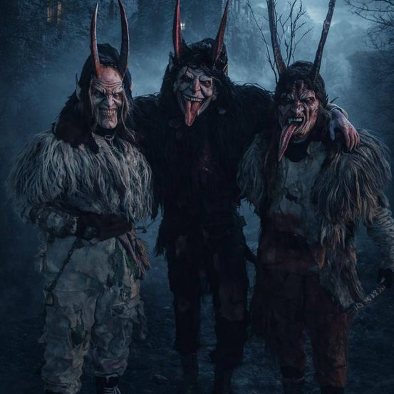 Drei Menschen in Pelzkostümen mit Hörnern und gruseligen Masken, die in einem dunklen Wald stehen. Die Person auf der linken Seite lächelt.