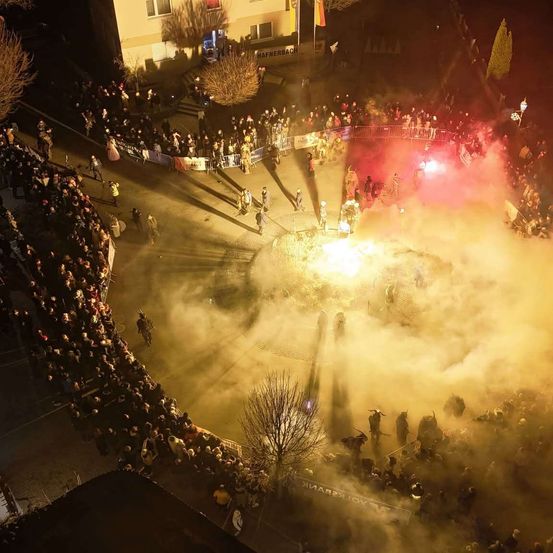 Eine oberirdische Ansicht eines überfüllten Platzes mit einem Feuer in der Mitte. Rauch und rote Lichter füllen die Luft, während sich Menschen versammeln und einige Kostüme tragen. Gebäude und Flaggen sind im Hintergrund zu sehen.