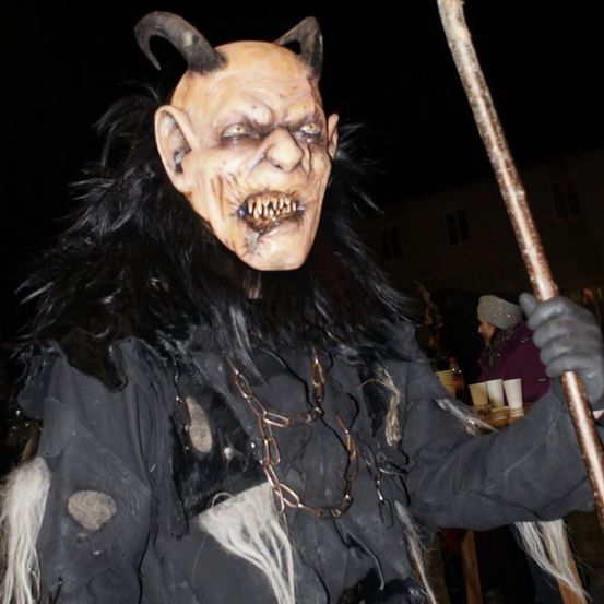 Eine Person in einem horntragenden Dämonenkostüm mit einem Stock, Ketten und einer gruseligen Maske, wahrscheinlich auf einer Halloween-Party.