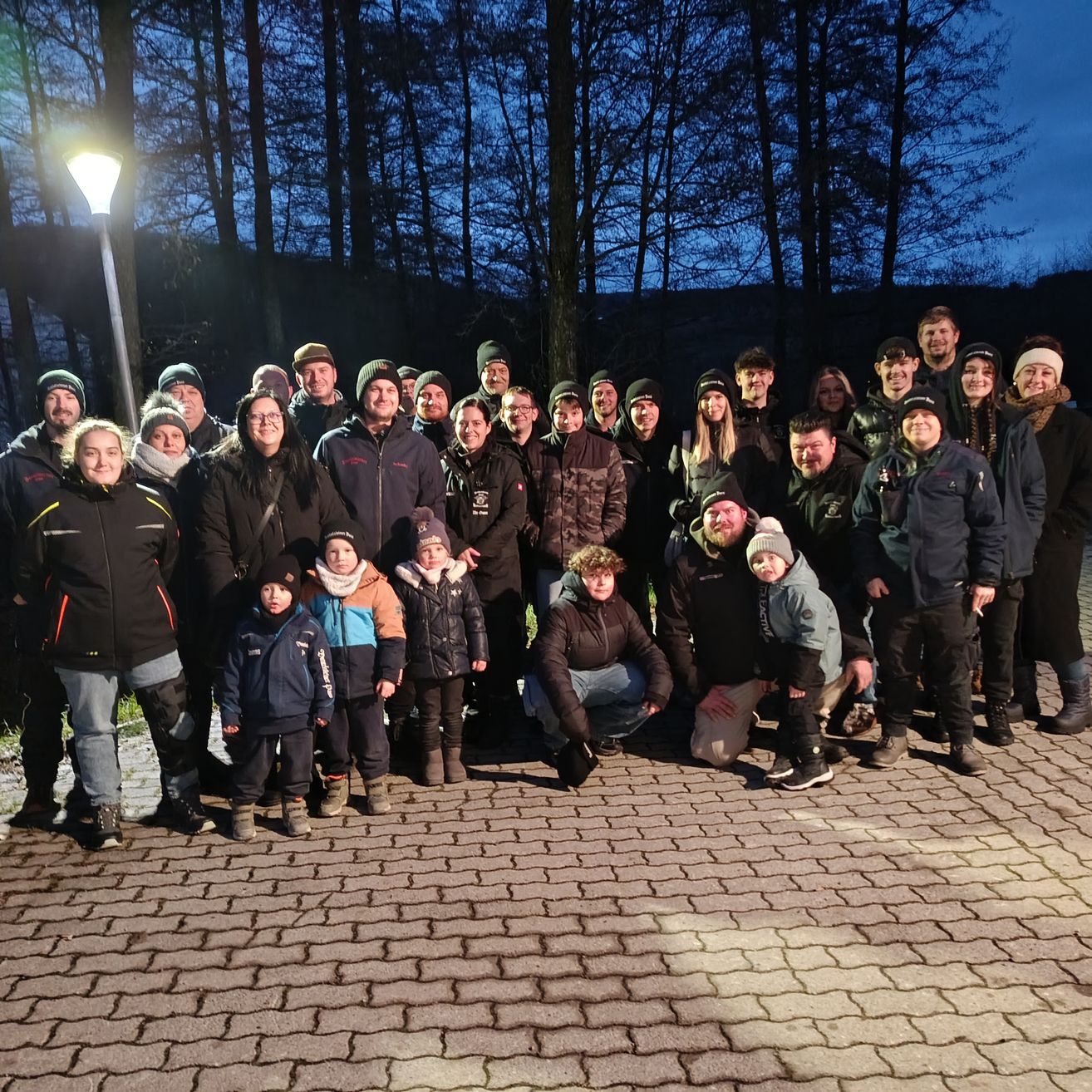 Eine Gruppe von Menschen in Winterkleidung, einige mit Mützen und Brillen, posieren für ein Foto auf einem Ziegelweg in einem Wald.