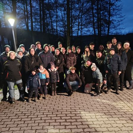Eine Gruppe von Menschen in Winterkleidung, einige mit Mützen und Brillen, posieren für ein Foto auf einem Ziegelweg in einem Wald.