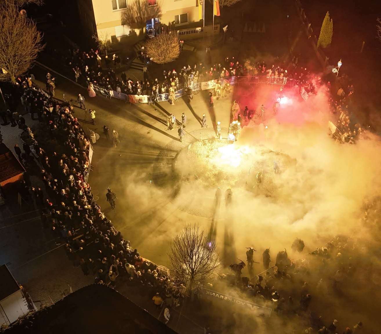 Eine nächtliche Menschenmenge versammelt sich um ein Feuer mit Rauch, wahrscheinlich bei einem Festival. Einige Leute tragen Kostüme. Gebäude säumen den Hintergrund, und Flaggen sind sichtbar.