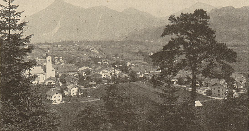 Ein Sepia-gefärbtes Foto eines Dorfes am Fuße der Berge. Ein Kirchturm ragt zentral heraus. Bäume und Häuser säumen die Landschaft.