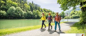 Drei Frauen wandern entlang eines malerischen Seepfades. Sie tragen Rucksäcke und Wanderschuhe, mit einem friedlichen Waldhintergrund. Der Text 'Barrierefrei geprüft' zeigt an, dass der Pfad barrierefrei ist.