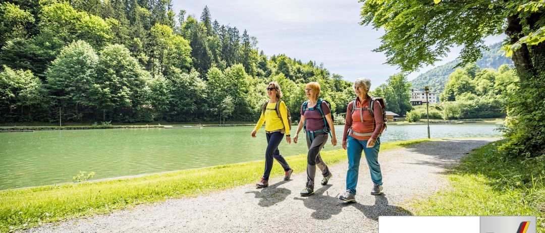 Drei Frauen wandern entlang eines malerischen Seepfades. Sie tragen Rucksäcke und Wanderschuhe, mit einem friedlichen Waldhintergrund. Der Text 'Barrierefrei geprüft' zeigt an, dass der Pfad barrierefrei ist.