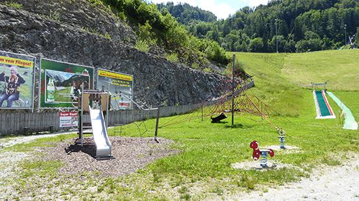 Freiluftspielplatz mit Rutsche und Klettergerüst nahe einer Felswand, mit Werbeplakaten an der Wand.