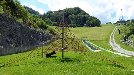 Ein Spielplatz mit einem Netzaufbau und einer Rutsche auf einem Grashügel. Im Hintergrund befinden sich Berge und ein Weg.