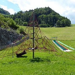 Ein Spielplatz mit einem Netzaufbau und einer Rutsche auf einem Grashügel. Im Hintergrund befinden sich Berge und ein Weg.