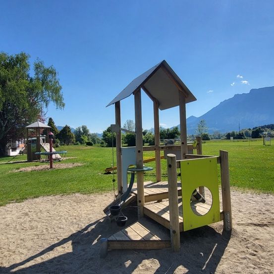 Eine hölzerne Spielstruktur befindet sich in einem sandigen Bereich mit grünem Gras. Sie verfügt über einen Kletterrahmen und eine Schaukel. In der Nähe befindet sich eine Rutsche. Im Hintergrund sind Berge zu sehen.
