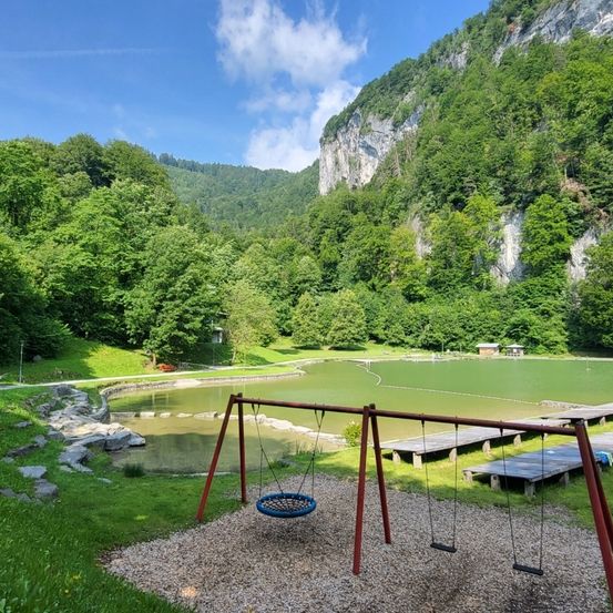 Ein ruhiger Außenspielplatz mit Schaukeln und einem kleinen Teich, umgeben von üppigem Grün und Bergen im Hintergrund.