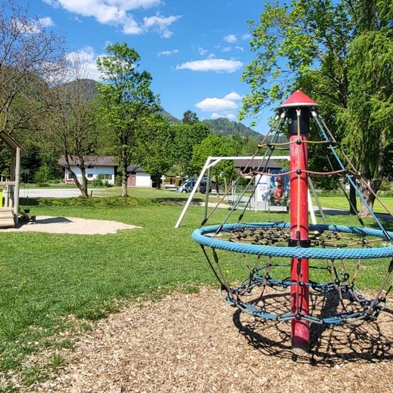 Ein Spielplatz mit Klettergerüst und Schaukel unter blauem Himmel, umgeben von Bäumen und einem Rasenfeld mit Häusern im Hintergrund.