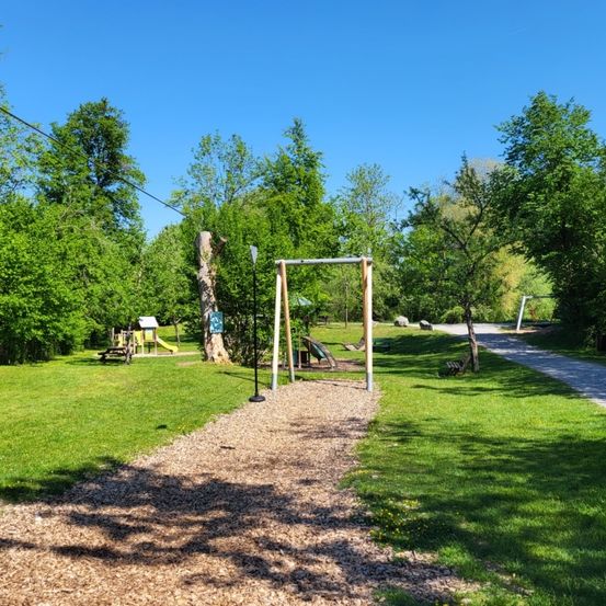 Ein Spielplatz mit Schaukel, Rutsche und Picknicktisch liegt unter einem klaren blauen Himmel. Bäume und ein Weg umgeben das Gelände.