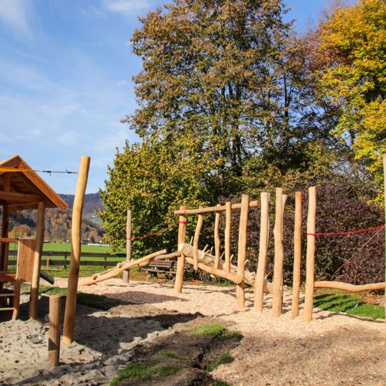Ein Outdoor-Spielplatz mit Holzstrukturen, einer Rutsche und einem kleinen Haus, umgeben von Bäumen und einem Feld.