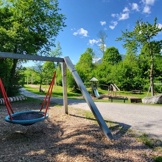 Ein Spielplatz-Szene mit Schaukel, Rutsche und Bänken unter einem klaren blauen Himmel mit Wolken, umgeben von üppigem Grün.