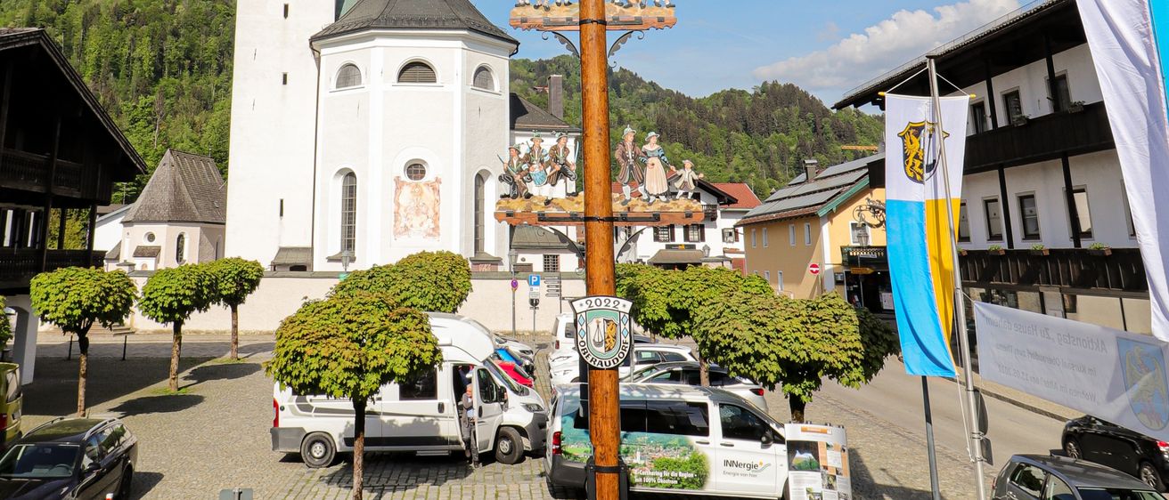 Ein großer verzierter Pfahl steht vor einer Kirche, umgeben von einer Gruppe von Bäumen und Fahnen. Die Kirche hat einen hohen Turm und einen klaren Himmel darüber.