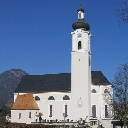 Eine weiße Kirche mit einem Kirchturm und Uhrenturm steht vor einer Bergkette. Das Gebäude hat ein schwarzes Dach und ein braunes Dach auf dem vorderen Teil.