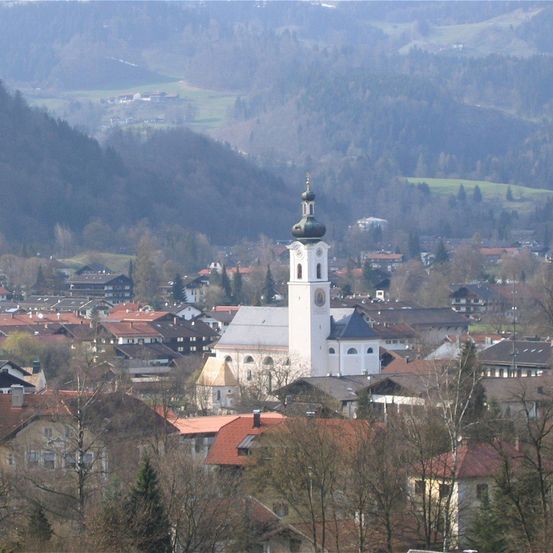Eine Stadt mit einer weißen Kirche und einer grünen Kuppel in der Mitte, umgeben von verschiedenen Gebäuden und Bäumen, mit Bergen in der Ferne.
