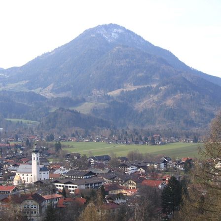 Ein Dorf in einem Tal unterhalb eines schneebedeckten Berges, mit einer Kirche und zahlreichen Gebäuden, umgeben von grünen Feldern und Bäumen.