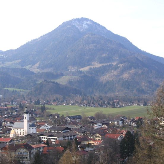 Ein Dorf in einem Tal unterhalb eines schneebedeckten Berges, mit einer Kirche und zahlreichen Gebäuden, umgeben von grünen Feldern und Bäumen.